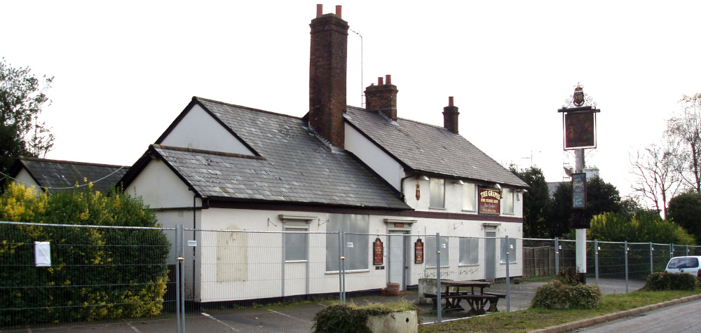An old white building with a grey tiled roof and a tall chimney, with a pub sign and a metal fence in front