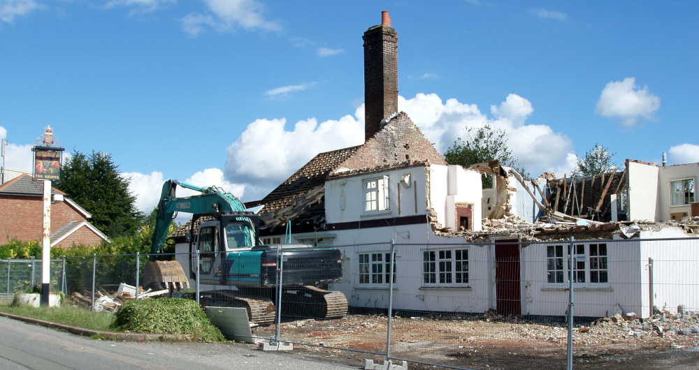 A partially destroyed white building with a pub sign outside, and a green mechanical digger