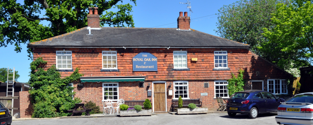 A wide, two-storey brick building with pantiles, with wooden benches in front and a sign on the wall reading: The Royal Oak Inn and Restaurant